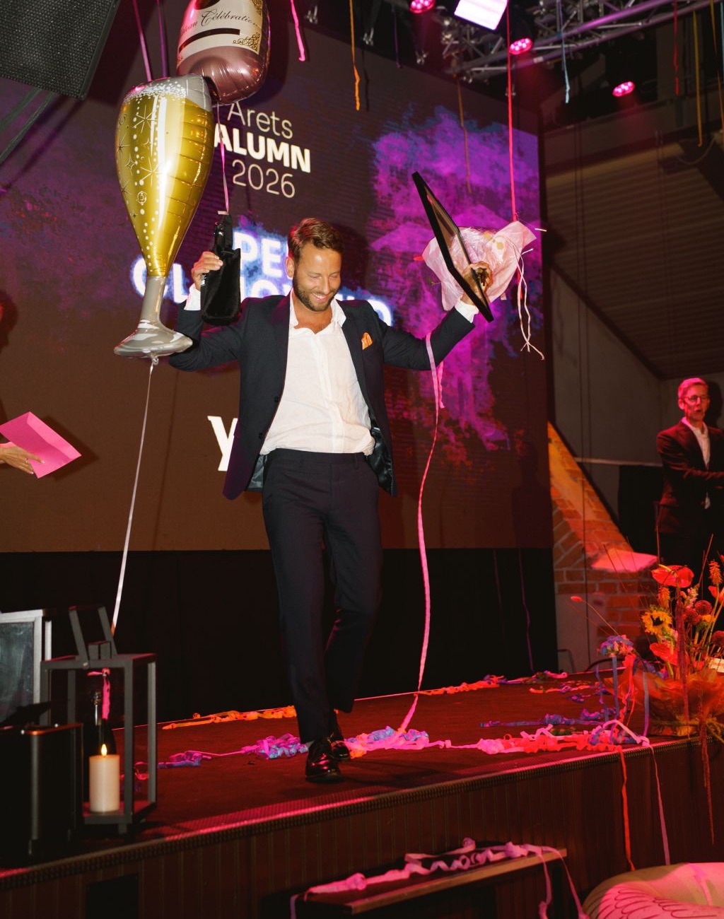 Per Clingweld går av scenen med diplom, blommor och ballonger framför skärmen Årets Alumn 2026